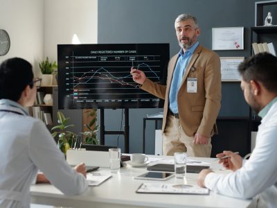 Waist up portrait of senior business expert presenting statistical data during medical conference and pointing at line graph on digital board while speaking to doctors