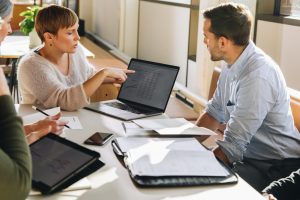 Manager pointing at laptop screen and talking to colleagues during business meeting. Female executive showing data to team in meeting.
