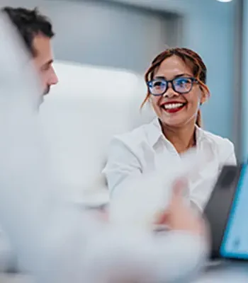 A smiling Filipino woman with blue glasses chats with a colleague in a modern office, laptop open showing charts. Warm, collaborative moment highlighting teamwork and communication.